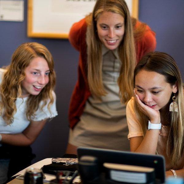 Three people, two of them sitting and one standing between them, smile while looking at a computer screen.