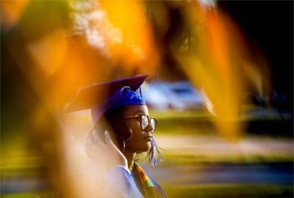   A student with a graduation cap and gown is pictured with orange and yellow autumn tree colors.