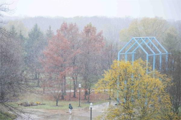  A pedestrian walks through a mix of snowy precipitation on campus filled with autumn tree colors.