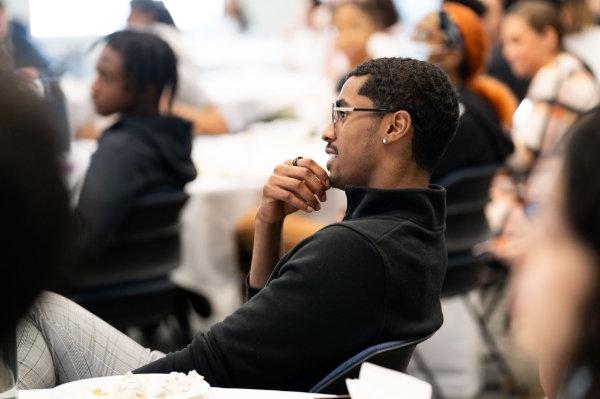 A person sits in a crowd touching their chin while listening to a presentation. 