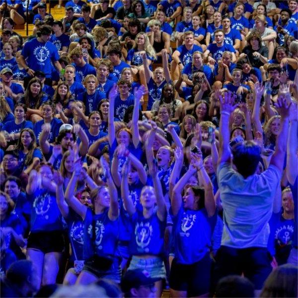 A group of students in blue Laker for a Life shirts sit in the bleachers and hold their arms up to do the wave.