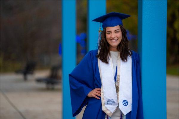 A person wearing graduation garb leans against a blue post.