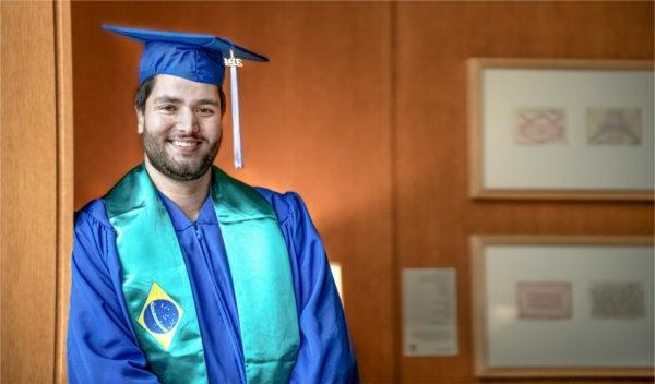 A person wearing academic garb smiles while leaning against a wall.