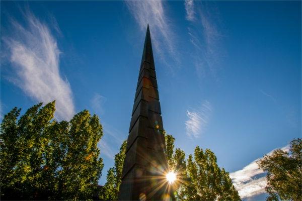  An obelisk sculpture juts into the blue sky as the sun peeks through. 