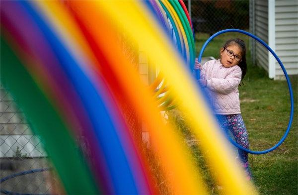  A small child wrestles with a blue Hula Hoop as they try to put it on a rack with a rainbow of colored hoops. 