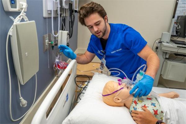  A nursing student wearing a blue scrub top puts an oxygen mask onto a simulation manikin. 