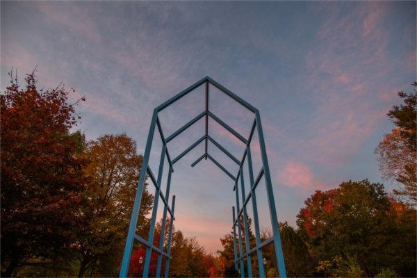  A blue metal sculpture is seen against a pink sunrise and colorful fall leaves. 