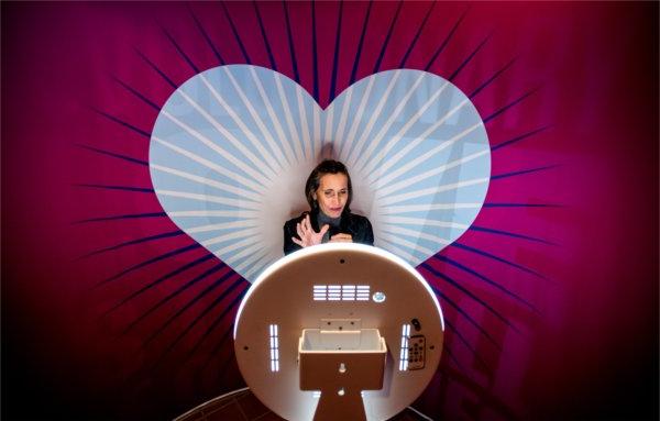  A person gestures while standing in front of a video recording screen with a large pink heart background. 