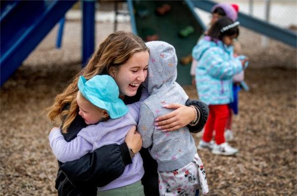  A college student hugs two small children while kneeling on a playground. 