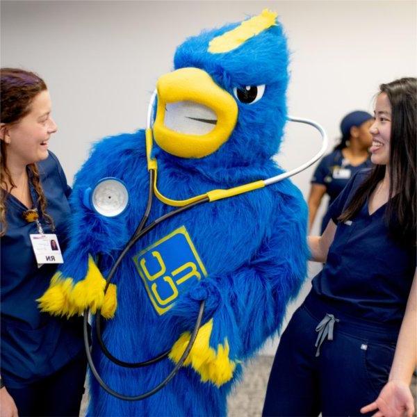 two nursing students in scrubs laugh at a Jaybird mascot with a stethoscope around its neck