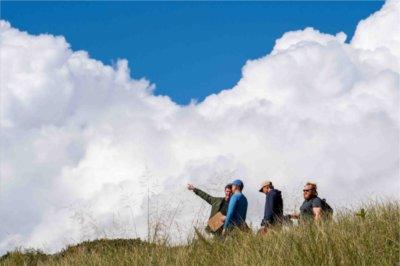 College students with their professor walk among dune grass as white puffy clouds loom behind them.