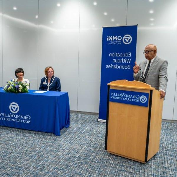 one person at podium in front of banner, two people seated at table with GV tablecloth