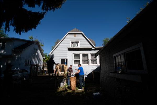 from the backyard, a two-story house in background, two people working in the yard talk to each other