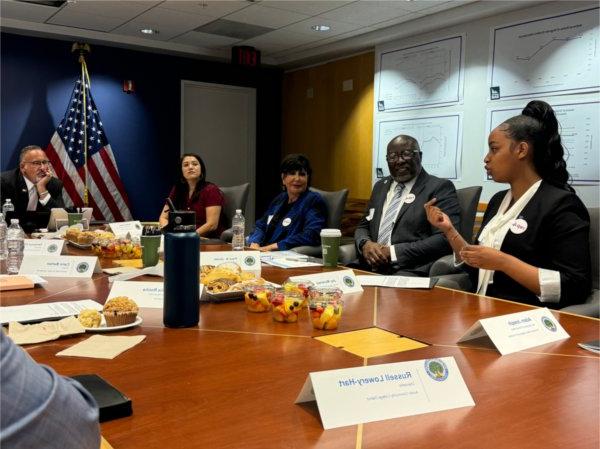 A group of people including Secretary of Education Miguel Cardona sit at a table and listen to a young woman, Joy Murerw speak.