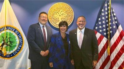 President Mantella stands between two men, an American flag flanking them on the left and the Department of Education flag on the right.