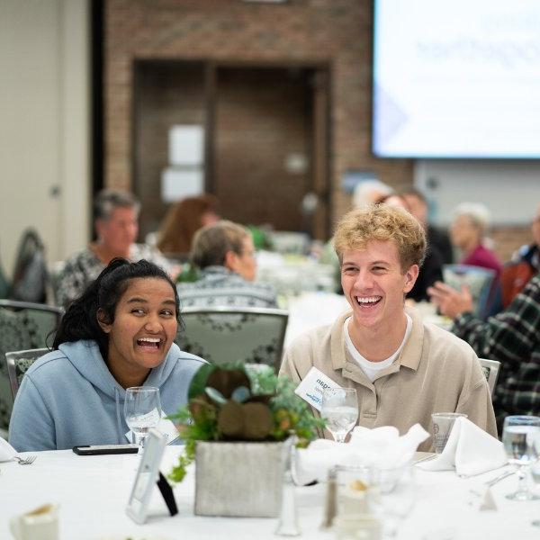 Participants at a Talking Together event in the Eberhard Center are pictured laughing, many tables with people seated behind them. Screen pulled down at front of room