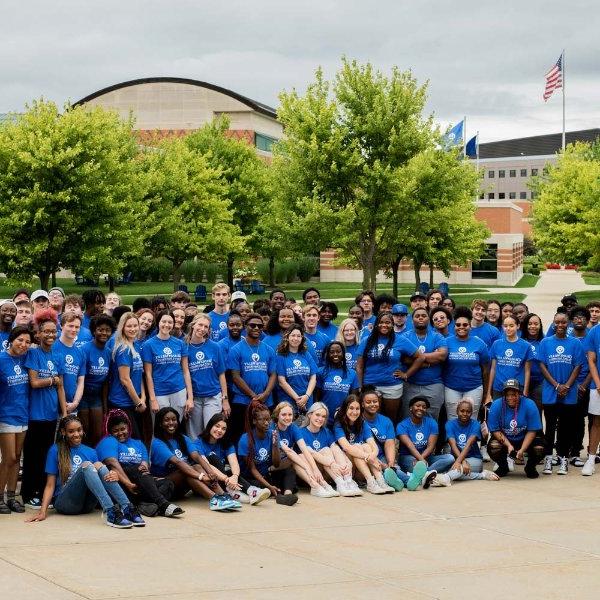 large group of students, faculty and staff all wearing blue GVSU Oliver Wilson Scholar t-shirts; pictured with Student Services Building behind them